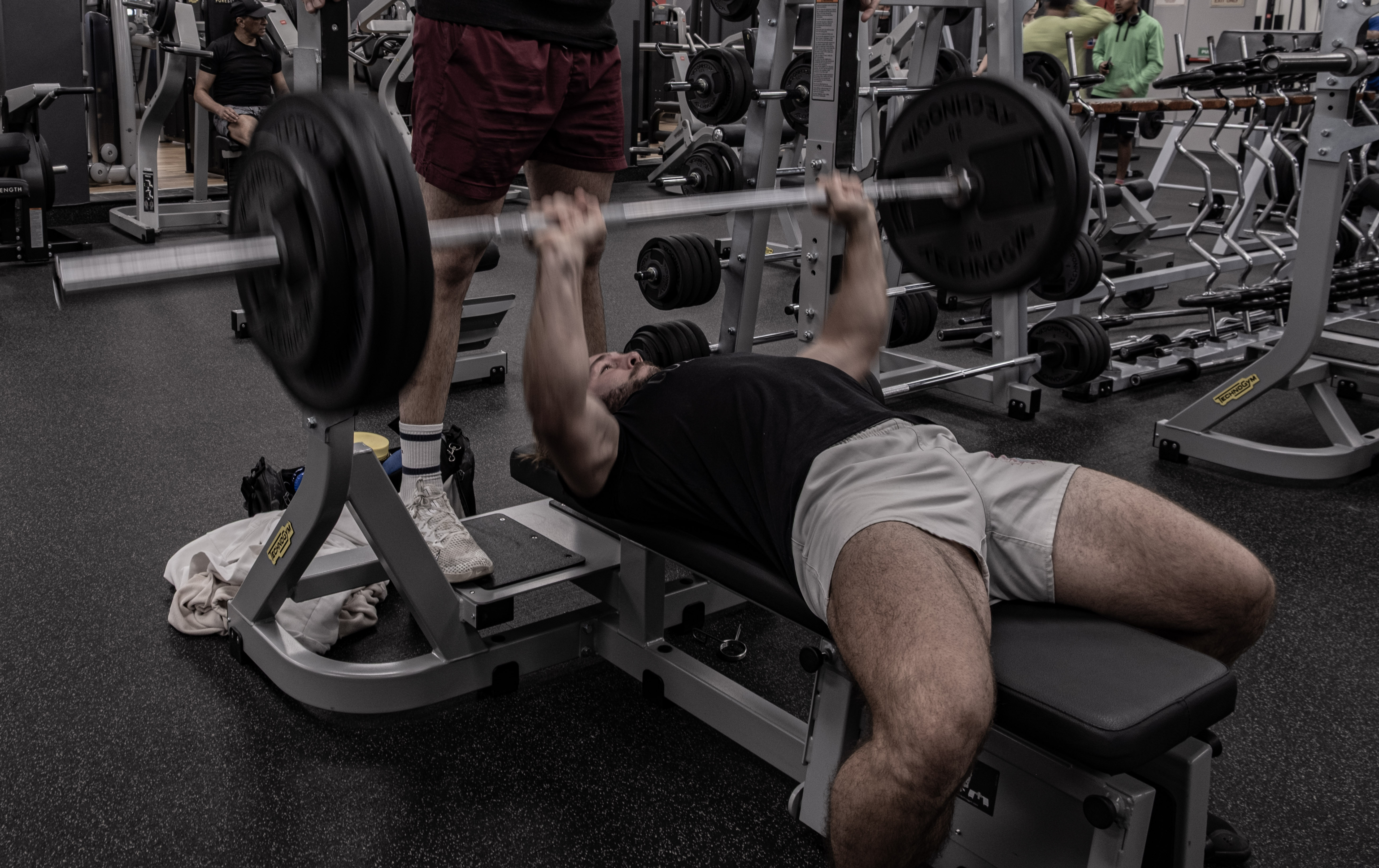 An athlete benching with a spotter standing on the bench platform, weight racks in the background.
